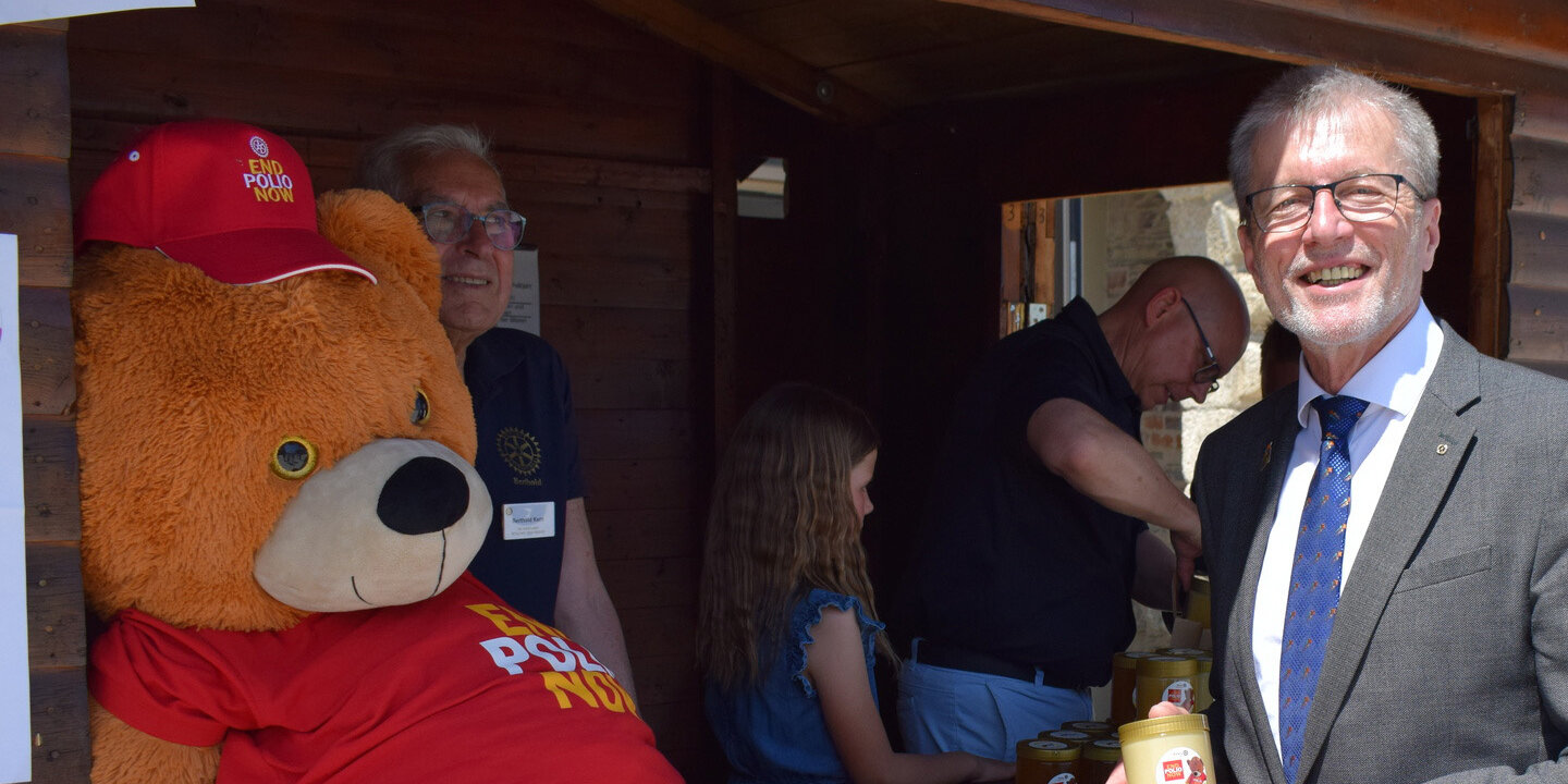 Pastgovernor Hans Neuser steht vor mit seinem Polio-Honig vor einer Hütte, die mit einen riesigen Polio-Bären geschmückt ist.